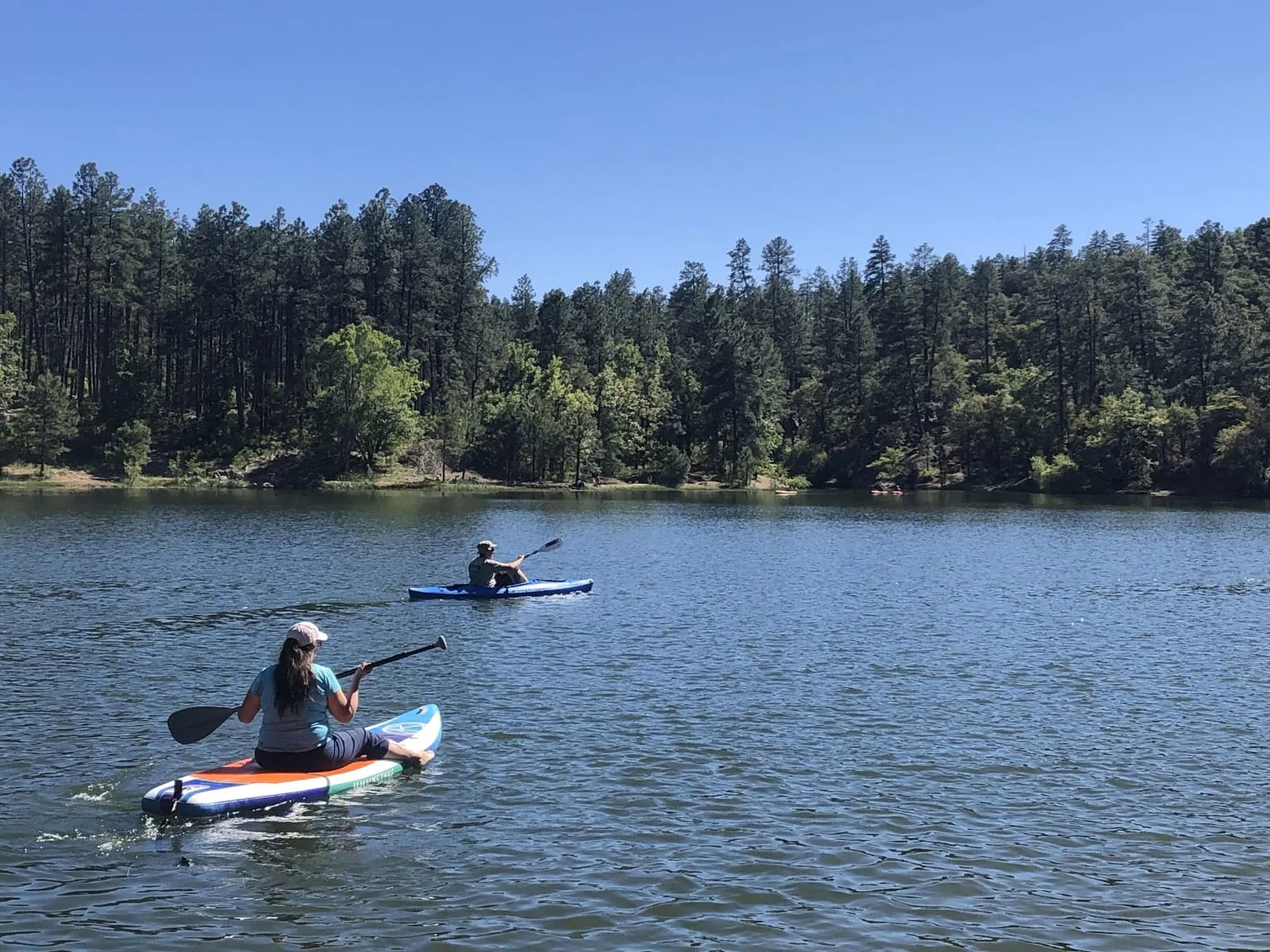 Two people paddling kayaks and a paddleboard on a calm, pine-ringed Prescott lake